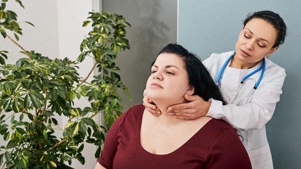 Healthcare professional stands behind woman patient who is sitting, and gently touches the front of the patient's throat.