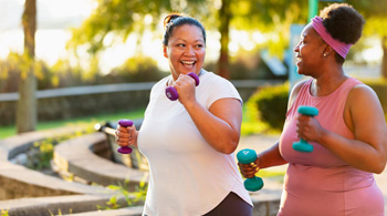 Two women walking throug a park in exercise clothes, laughing while carrying hand weights.