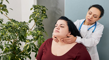 Healthcare professional stands behind woman patient who is sitting, and gently touches the front of the patient's throat.