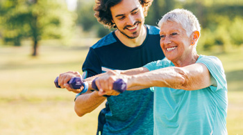 Older adult working out with light dumbells while outside with a personal trainer.