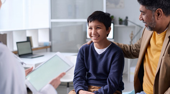 Father holding on adolescent son's shoulder while listening to healthcare provider in an healthcare setting.